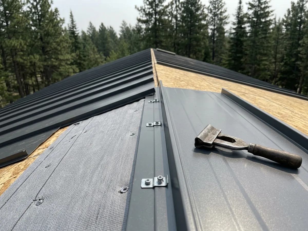 Charcoal standing seam metal panels being installed over synthetic underlayment, with concealed clips along the seam and a hand seamer resting on a panel